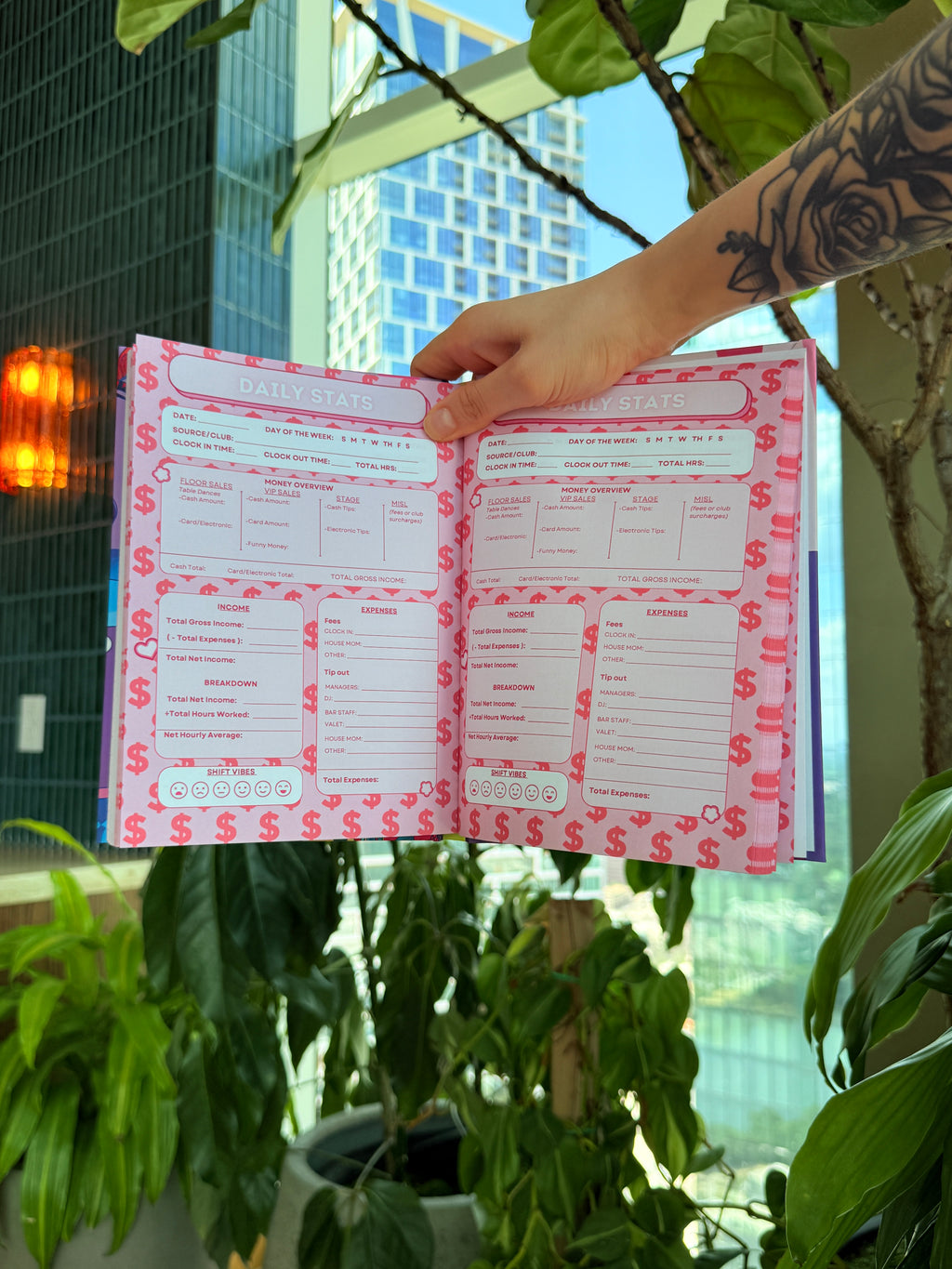 A hand with a floral tattoo holds an open pink budget planner or notebook with dollar-sign patterned pages, set against a background of green potted plants and a city building.