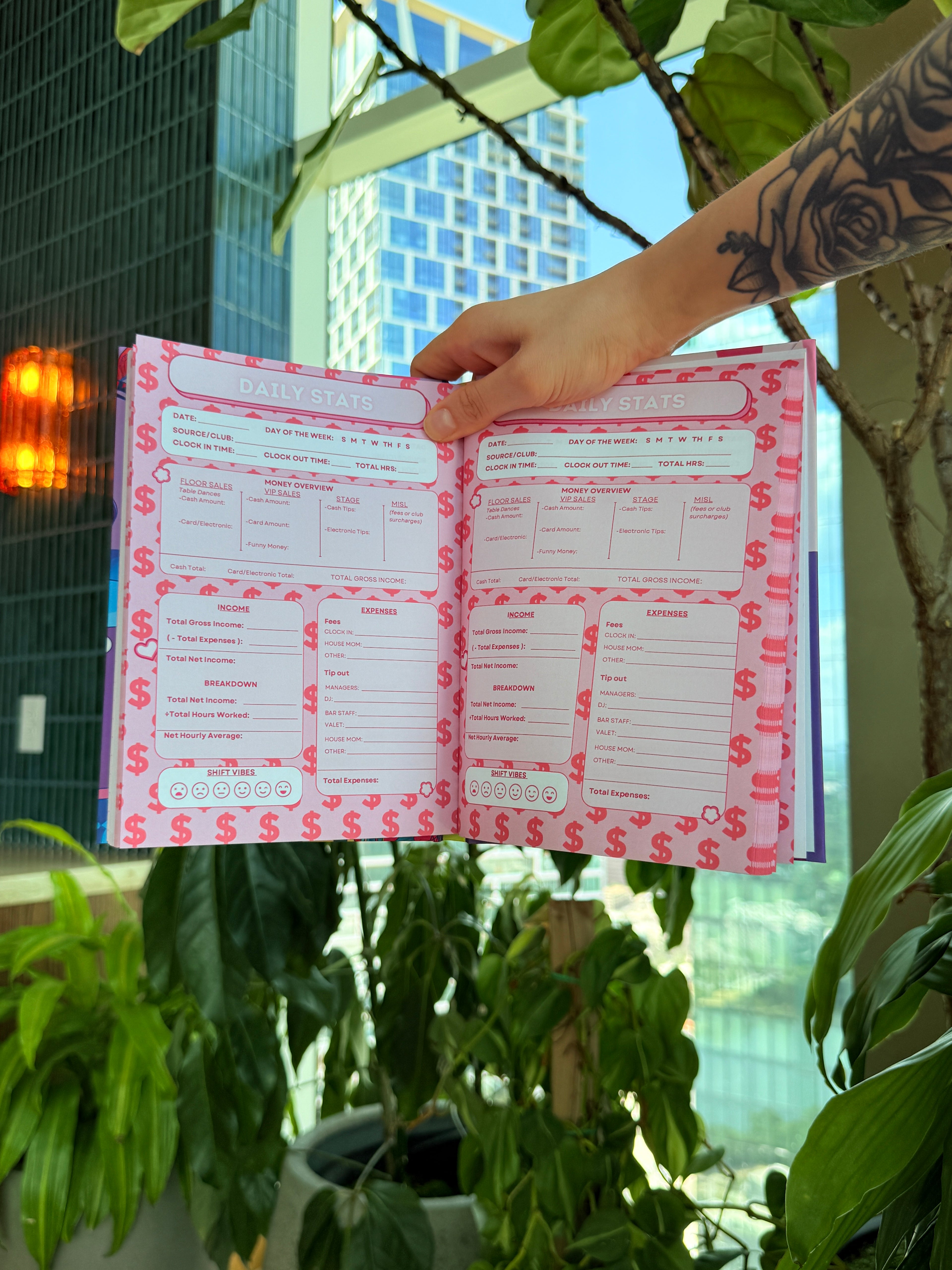 A hand with a floral tattoo holds an open pink budget planner or notebook with dollar-sign patterned pages, set against a background of green potted plants and a city building.