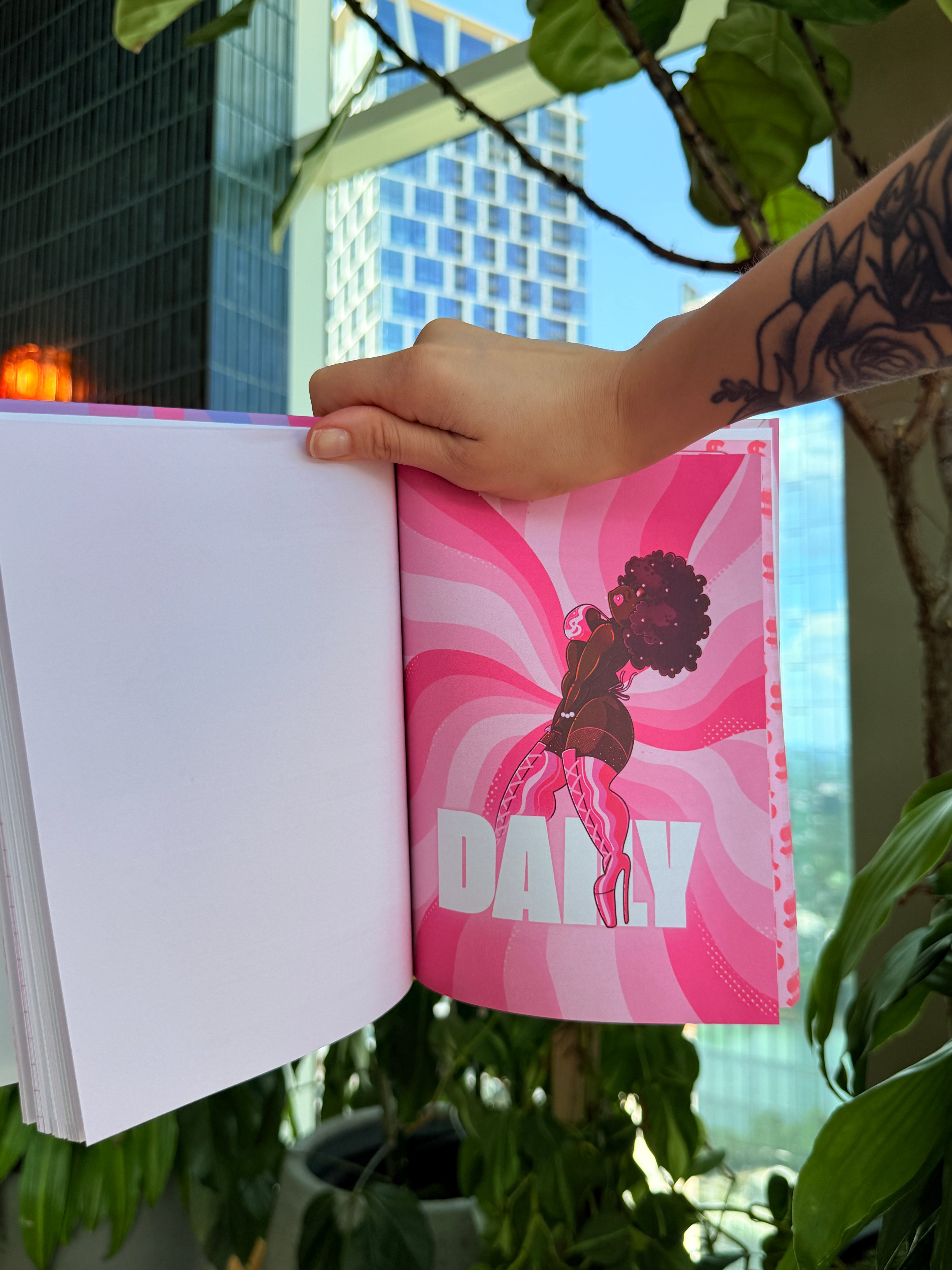 A hand with a floral tattoo holds open a book or magazine showing a bright pink illustrated page with the word 'DAILY' on the right page, with a real-world background of green plants and a city building.