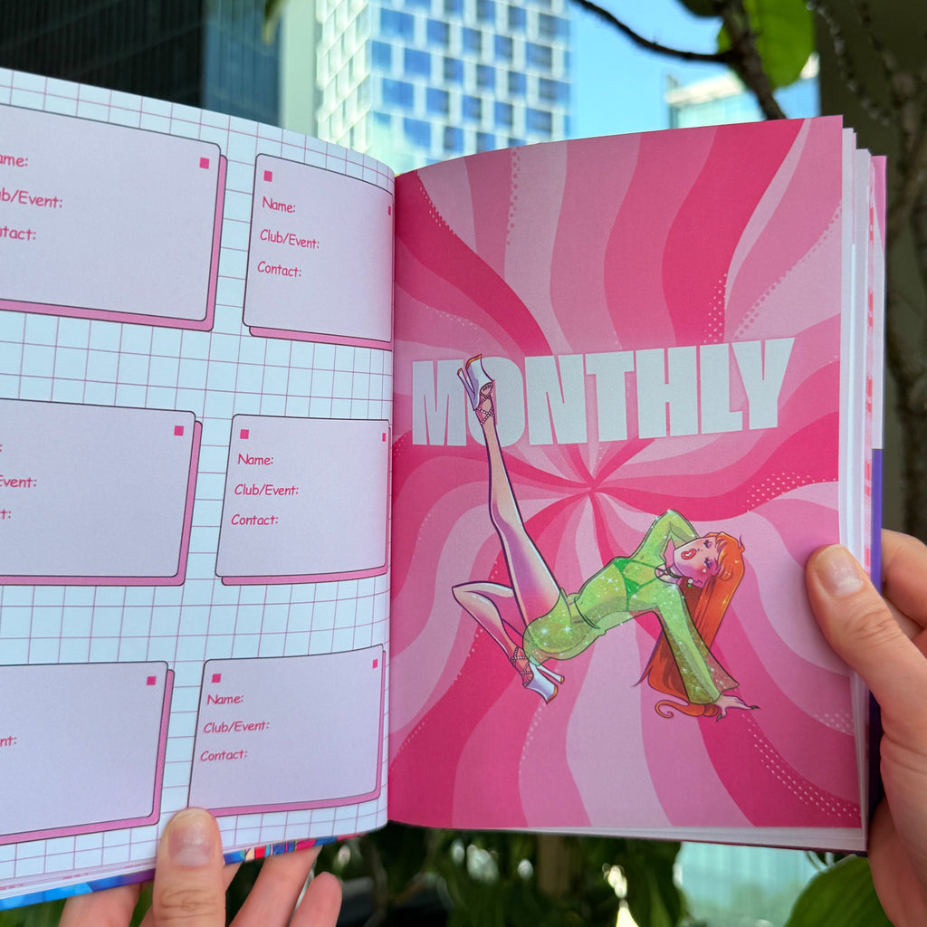 A person holding open a pink monthly planner or journal, showing a colorful cover with a cartoon figure and a grid layout for daily entries, photographed outdoors with plants and a building visible in the background.
